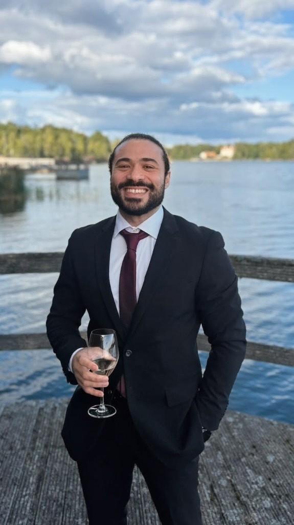 Aly Metwaly in a formal suit, smiling on a wooden dock beside the water.